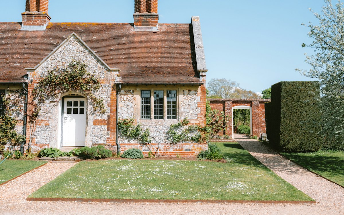 Photo of the cottage from the lane: a flint-and-brick front with a white door, climbing plants, two chimneys and a lawn.